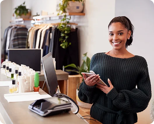 Smiling boutique owner holding a phone beside a point-of-sale terminal, cybersecurity for retail businesses