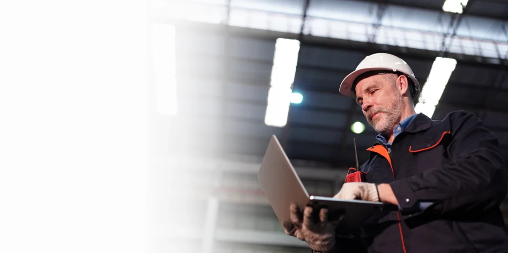 Plant engineer in a hard hat checking a laptop in an industrial facility, OT ICS cybersecurity