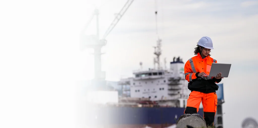 Port engineer in hi-vis safety gear using a laptop beside a cargo ship for maritime cybersecurity
