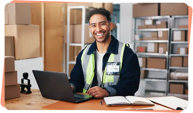 Smiling warehouse worker in a hi-vis vest using a laptop among shipping boxes